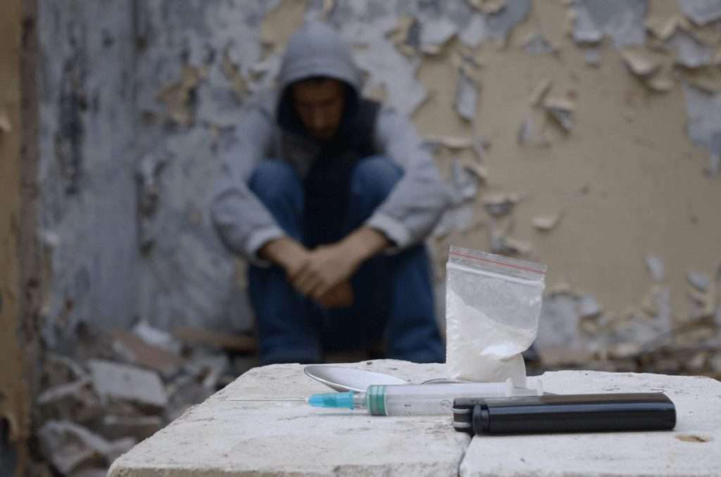 Un hombre encapuchado sentado contra una pared junto a una mesa con drogas y una jeringa