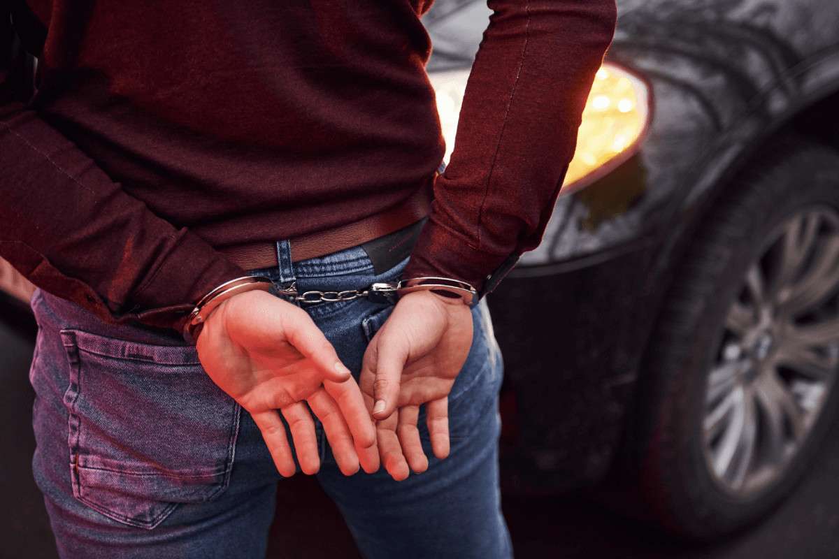 Un hombre esposado a la espalda junto a un coche con los faros encendidos